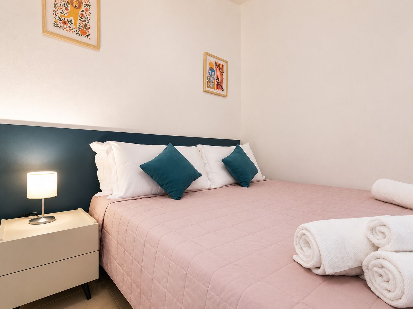 A hotel room with a bed, two pillows, a bedside lamp, a nightstand, and a rolled towel.