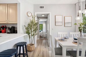 A modern kitchen with wooden cabinets, a dining area with a white table and chairs, and a hallway leading to a bedroom. - Renovated 2BR Condo Walk to Beach Pool Decks (Hilton Head Island)