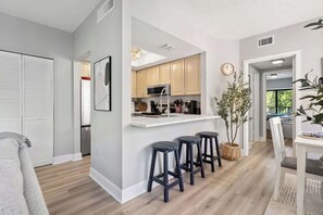 A modern kitchen with a white countertop, wooden cabinets, and a microwave. There are three stools and a potted plant. - Renovated 2BR Condo Walk to Beach Pool Decks (Hilton Head Island)