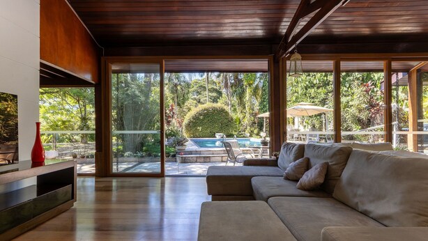 Una sala de estar moderna con un sofá grande, un televisor y vistas a una piscina y un jardín a través de grandes puertas corredizas de vidrio.