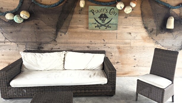 A wicker sofa with cushions, a wicker chair, and a wooden wall with a sign reading 'Pirate's Cove.' - Capn's Corner--1 BR, 1 Bath, Garage, Close to Beach, Dogs OK! (Port Aransas)
