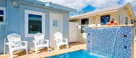 A swimming pool with two white chairs, a tiled pool area, and a building with a clock and a lifebuoy.