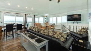 A spacious living area with a high ceiling, a ceiling fan, a dining table with chairs, a sofa set, a television, and large windows offering a view of the ocean. - Direct Beach Access Classic Surfside Home (Surfside Beach)