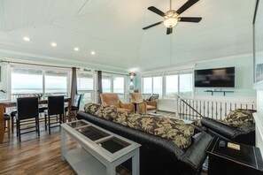 A spacious living area with a high ceiling, a ceiling fan, a dining table with chairs, a sofa set, a television, and large windows offering a view of the ocean. - Classic Surfside Beach Home with Ocean Views (Surfside Beach)