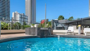 En swimmingpool med liggestole, en flisebelagt terrasse og høje bygninger i baggrunden.