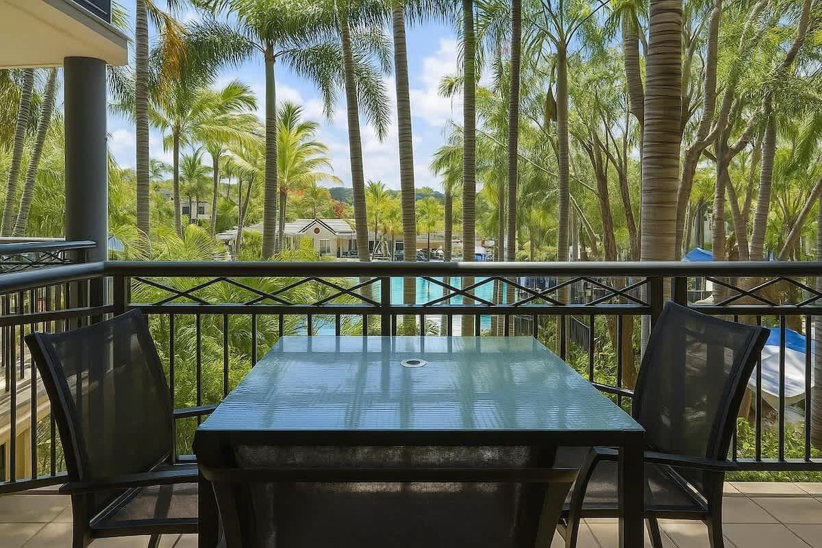 Un balcon avec une table et des chaises, donnant sur une piscine et des palmiers.