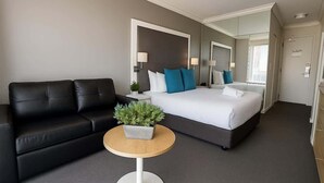 Ein Hotelzimmer mit schwarzem Ledersofa, einem Bett mit blau-weißen Kissen, einem runden Holztisch mit Pflanze und einem großen Spiegel über dem Bett.