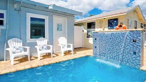 A swimming pool with a tiled edge, white chairs, and a building with a blue exterior.