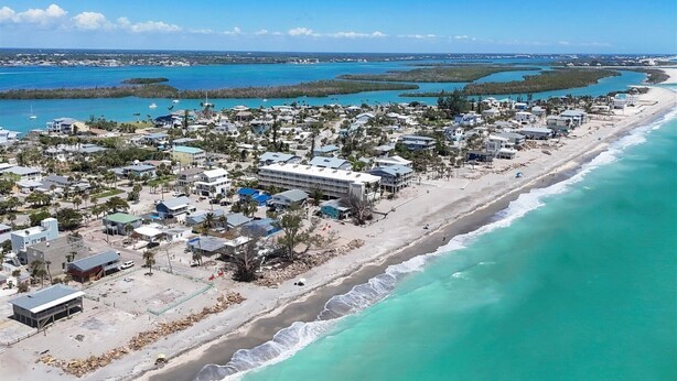 An aerial view of a coastal town with beachfront properties, a marina, and a clear blue sky.