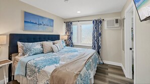 A bedroom with a bed, a painting, a window with curtains, an air conditioning unit, and a door.