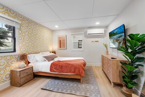 Een slaapkamer met een bed, nachtkastje, kaptafel, groot raam, decoratieve wandkunst en een potplant.