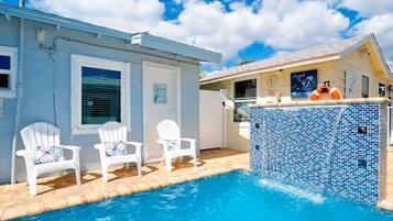 A swimming pool with two white chairs, a tiled pool area, and a building with a clock and a lifebuoy.