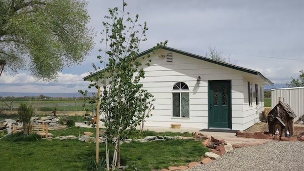 A small, single-story white building with a green door and windows, surrounded by a grassy area and a gravel path.