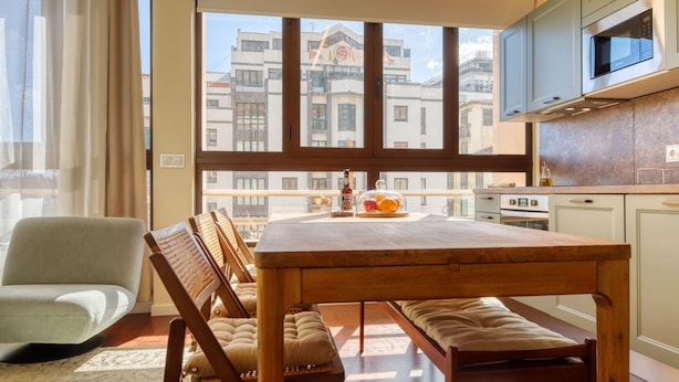 A dining area with a wooden table and benches, a kitchen with white cabinets, and a view of buildings through a large window. - Refuge in Funchal II, a Home in Madeira (Funchal)