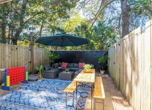 A backyard with a wooden fence, a blue patterned rug, a wooden table, a bench, a sofa, and an umbrella. - Wonderful 2-bedroom townhome close to beach +Pool (Myrtle Beach)