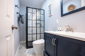 Ein Badezimmer mit Toilette in Weiß, einer Duschkabine aus Glas, einem dunkelblauen Waschtisch mit weißer Arbeitsplatte und einem gerahmten Bild an der Wand.