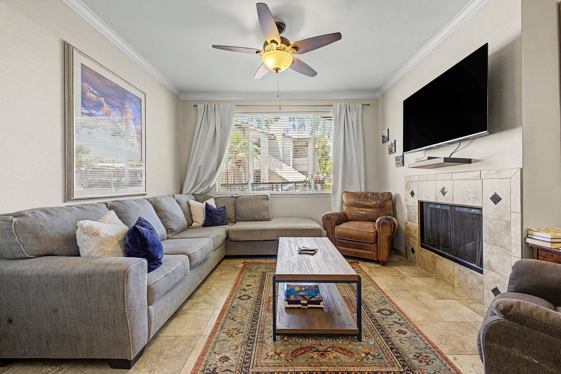 A living room with a sofa, armchair, coffee table, fireplace, TV, ceiling fan, and a large window with blinds.