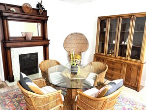 Ein Zimmer mit einem Glastisch, umgeben von Korbstühlen, einem Holzschrank mit Glastüren und einem Kamin, über dem sich ein Spiegel befindet.