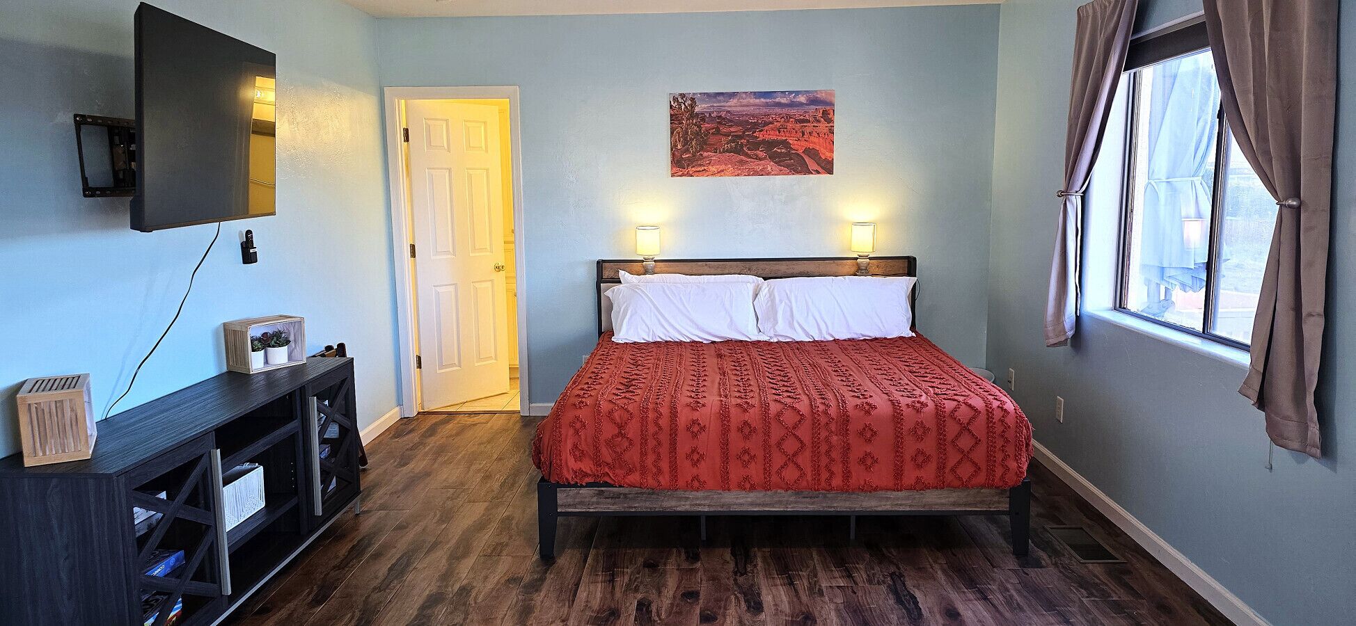 Un dormitorio con cama, televisor montado en la pared, una puerta, ventana con cortinas y un cuadro en la pared.