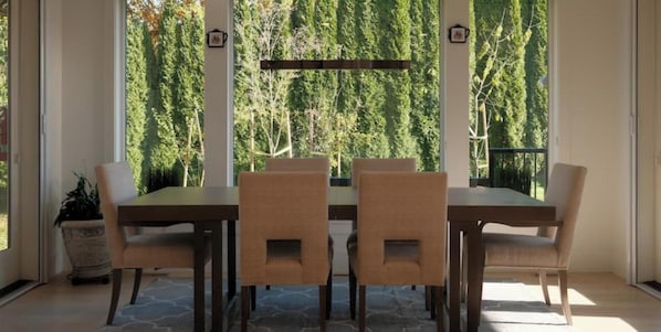 Un comedor con una mesa y sillas, ventanas amplias con vistas a árboles y una planta en maceta.
