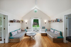 A room with a high ceiling, a central table, two wicker sofas with patterned cushions, and bookshelves with books.