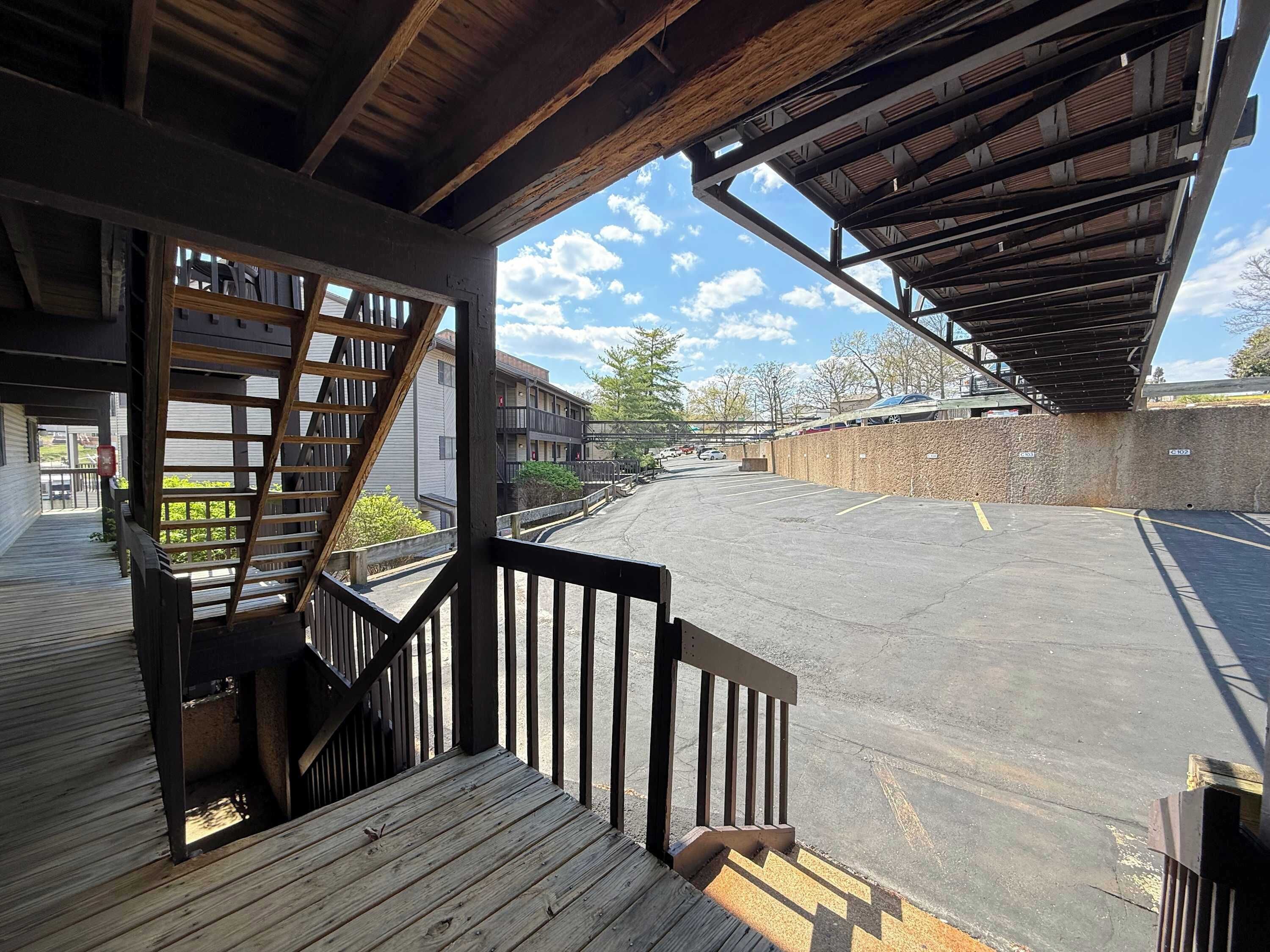 Un espace extérieur abrité, doté d’une terrasse en bois, d’un escalier et offrant une vue sur un parking et des bâtiments en arrière-plan.