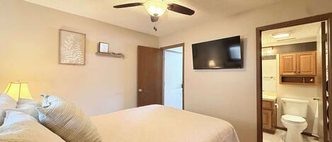 A bedroom with a bed, a ceiling fan, a TV, and a door leading to a bathroom.