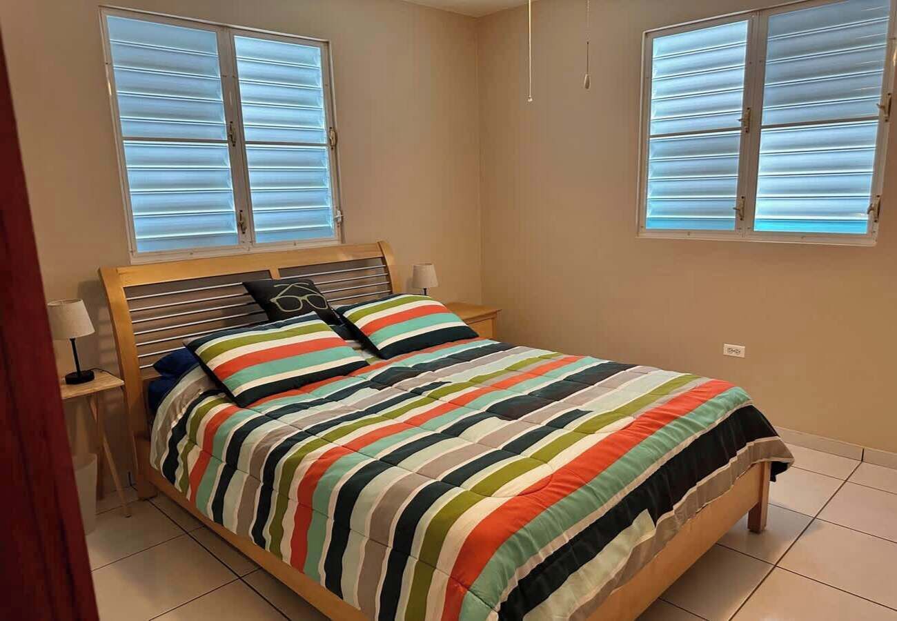 Un dormitorio con cama de madera, ropa de cama a rayas, dos mesitas de noche y una ventana con persianas.