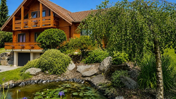 Een houten chalet met balkon, omgeven door een vijver met waterlelies en diverse struiken en bomen.
