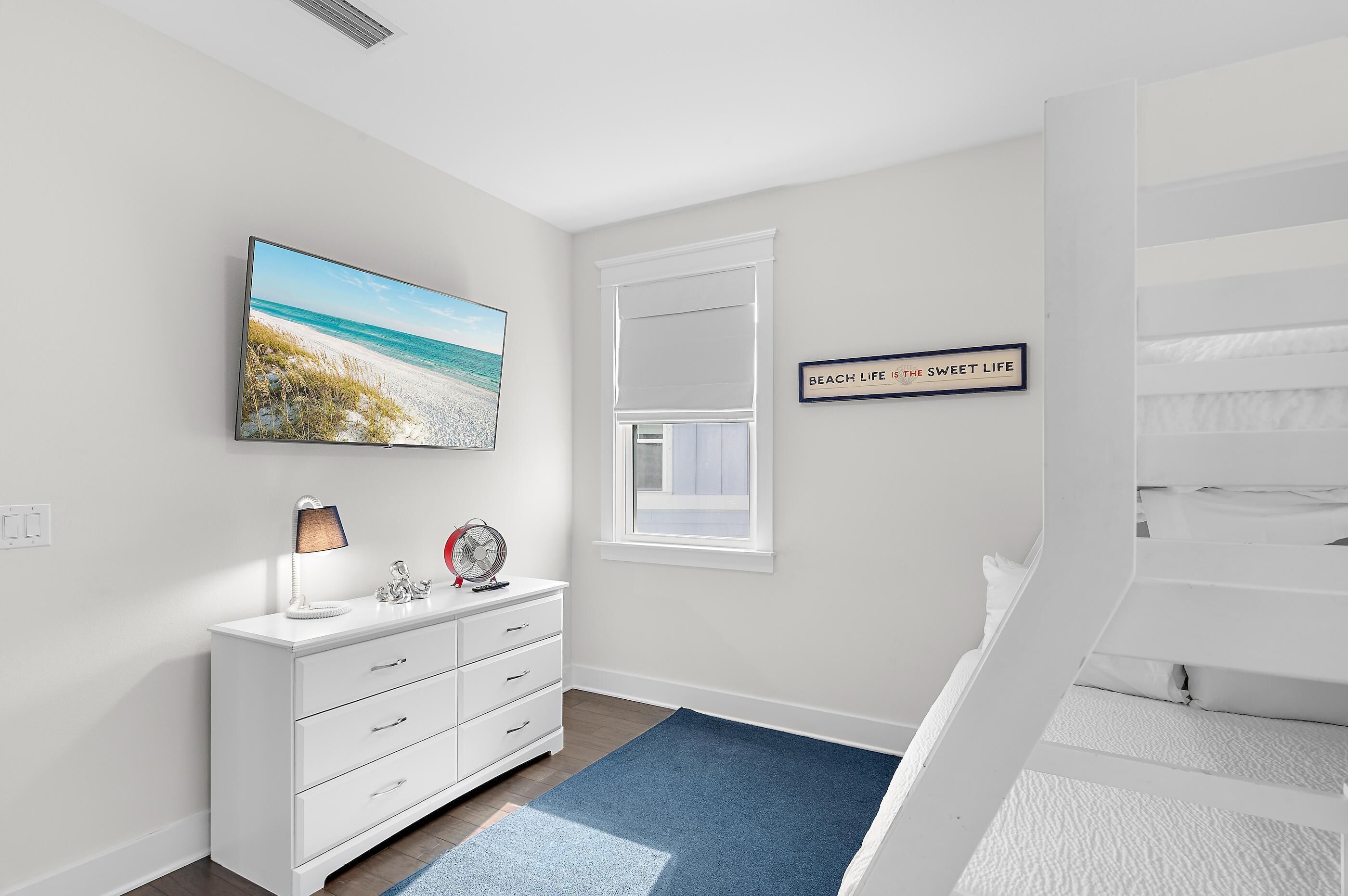 Une chambre avec un lit, une commode, une fenêtre avec des stores, un tableau sur le thème de la plage et un panneau accroché au mur.