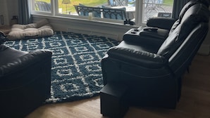 Una sala de estar con un ventanal grande, un sillón reclinable de cuero negro, un tapete azul con estampado y piso de madera.