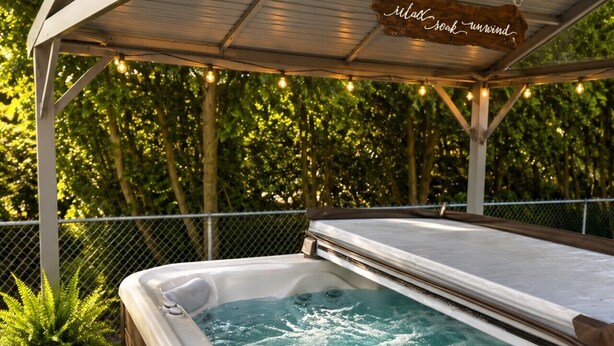 Un bain à remous sur une terrasse en bois, surmontée d’un toit en métal, le tout entouré de verdure.