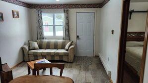 A small, cozy room with a sofa, a chair, a wooden table, a door, and a window with curtains. - Crocker Cabins Unit 1 offers a self‑contained efficiency unit in Gros Morne. (Trout River)