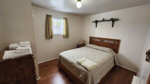 A bedroom with a wooden bed, a nightstand, a window with curtains, and a shelf above the bed. - Crocker Cabins Unit 1 offers a self‑contained efficiency unit in Gros Morne. (Trout River)