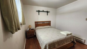 A bedroom with a wooden bed, a nightstand, a lamp, a shelf with decorative items, and a window with curtains.