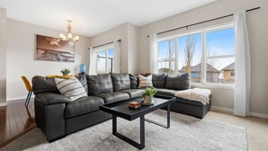 A modern living room with a sectional sofa, a coffee table, and a large window with a view of houses. - Merlot | AC | Crib | Fenced Yard | Pets Ok (Calgary)