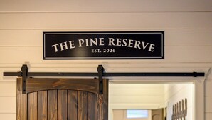 A hotel room with a wooden door labeled 'THE PINE RESERVE EST. 2026' and a view into the hallway with towels on a rack. - Luxury Resort | Golf Sim, Pool, Gym, Sauna, 30 ppl (Broken Bow)