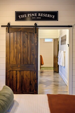 A hotel room with a wooden door labeled 'THE PINE RESERVE EST. 2026' and a view into the hallway with towels on a rack.