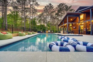 A swimming pool with lounge chairs and inflatable rings, surrounded by trees and a building with large windows.