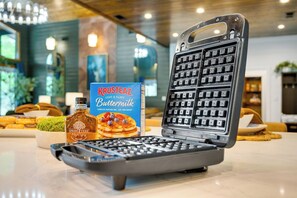 A waffle maker with a Krusteaz Buttermilk pancake mix box, a bottle of maple syrup, and a remote control on a table. - Luxury Resort | Golf Sim, Pool, Gym, Sauna, 30 ppl (Broken Bow)