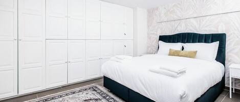 Quarto com cama, guarda-roupa branco, tapete estampado e parede com padrão de folhas.