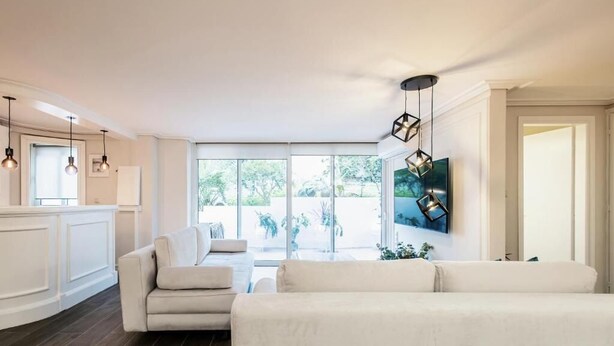 A modern living room with a sofa, a TV mounted on the wall, pendant lights, and a view of the outdoors through large windows. - MZ Kolonaki Hills (Athens)
