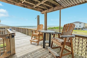 A wooden deck with two Adirondack chairs and a table, overlooking a beach and a building.