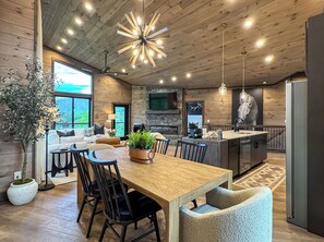 A modern kitchen with a wooden ceiling, a dining area with a large table and chairs, and a living room with a sofa and a fireplace.