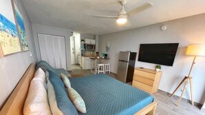 Een slaapkamer met een bed, kussens, een televisie, een bureau en een kitchenette op de achtergrond.