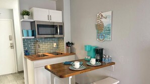 Een kleine keuken met witte kasten, een roestvrijstalen magnetron, een blauwe tegelachtergrond en een houten tafel met twee witte krukken.