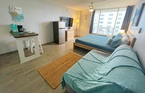 Une chambre de taille modeste, comprenant un lit, un petit bureau avec une chaise, un réfrigérateur, une télévision et une fenêtre donnant sur des immeubles.
