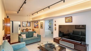 A modern living room with a sofa, coffee table, and TV. There is a dining area with a table and chairs. The room has a high ceiling with track lighting. - Leme 300 m praia 2 quartos conforto (Rio de Janeiro)