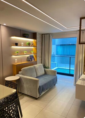 A modern living room with a sofa, a small table, a decorative shelf with plants, and a view of a blue balcony. - Altamare 110 - Luxurious beachfront apartment for 6 people. (João Pessoa)