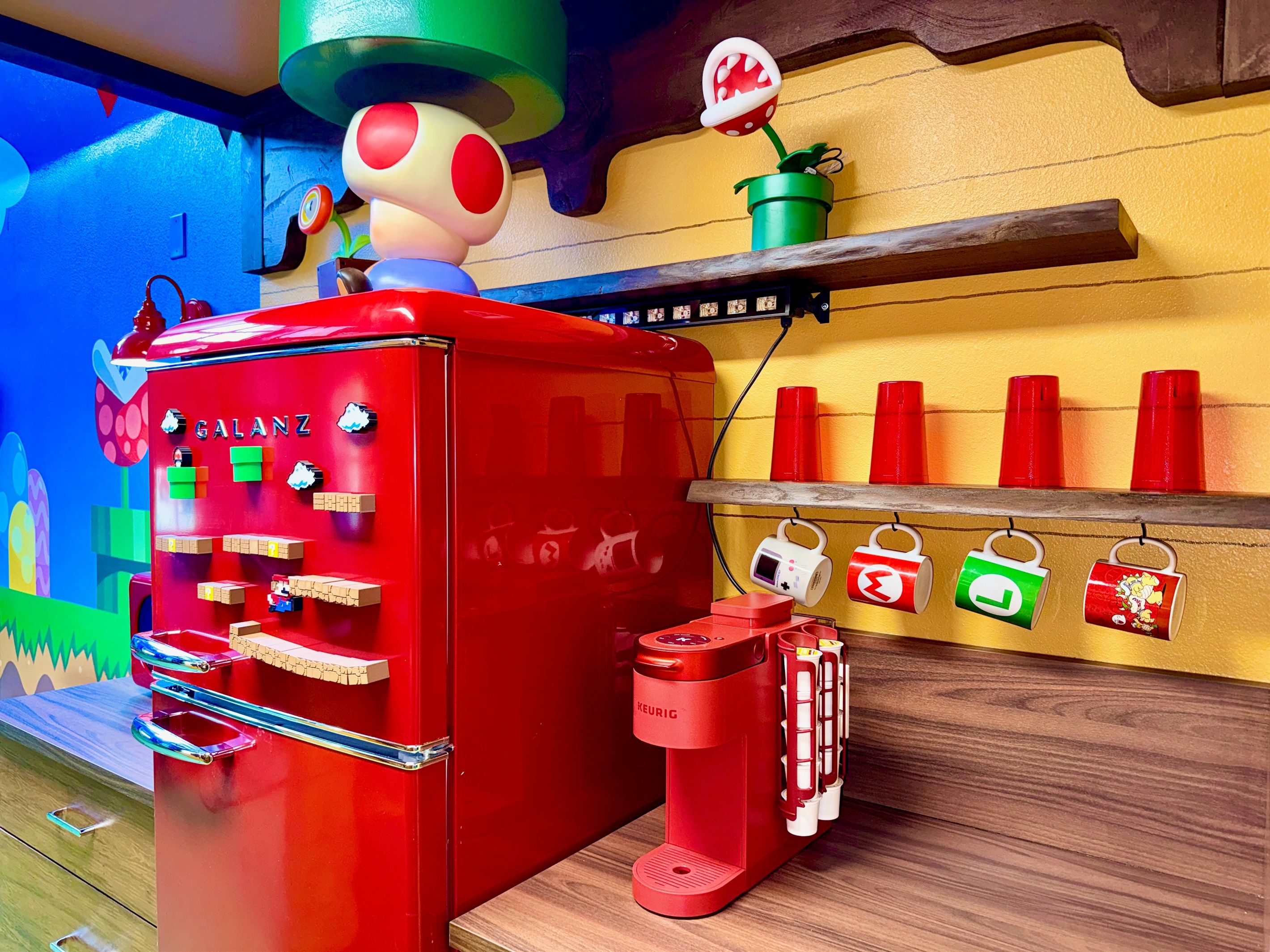 Ein roter Kühlschrank mit einer Kaffeemaschine, ein Regal mit roten Tassen und eine Wand mit einer Pilzdekoration.